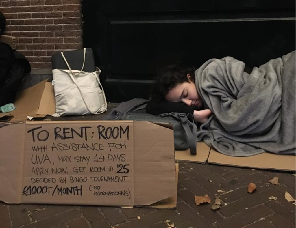 a student acting as if they are homeless and lying on cardboards with a sign titled "To rent: Room"